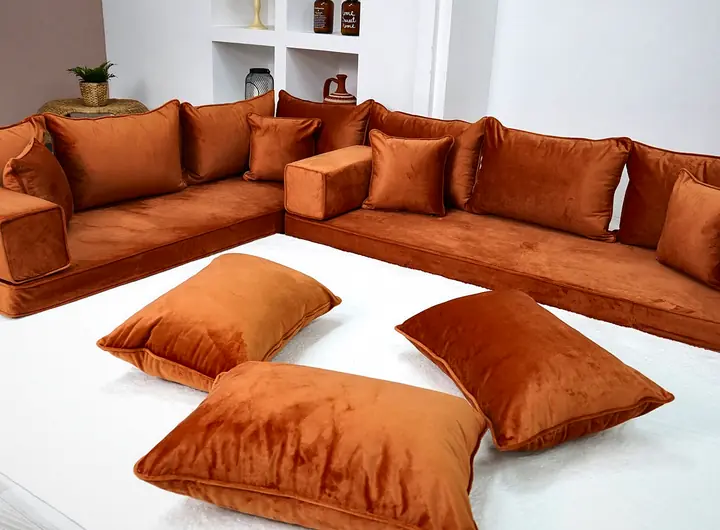Amber velvet L-shaped floor couch with plain backrests and three loose floor pillows set on a white rug in a bright room