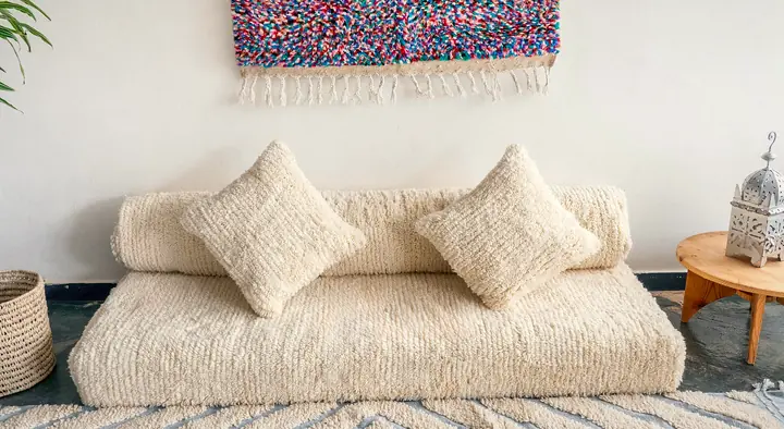 Cream shaggy floor couch with a long round bolster and two square pillows below a colorful woven wall hanging