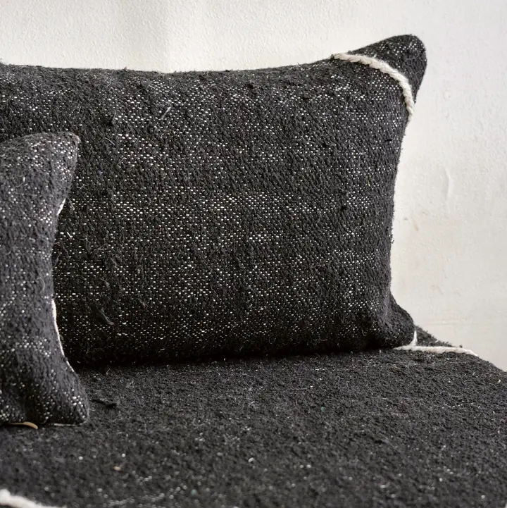 Close view of a charcoal black woven pillow and seat cover with thin white piping along the edges, against a white wall