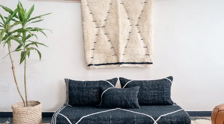 Wide view of a black lozenge-pattern floor couch with a wall hanging, a potted plant and a leather pouf nearby