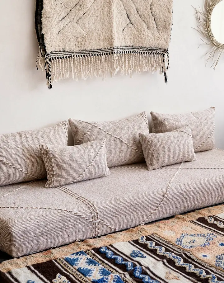 Beige Moroccan-style floor sofa with stitched lines and three long backrests, a striped rug, a woven wall hanging and a round mirror