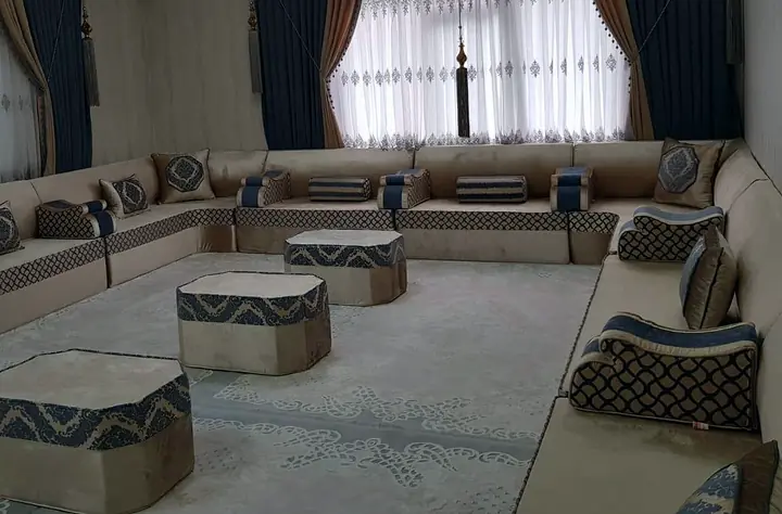 Beige velvet U-shaped majlis with navy wave-pattern bands running under two wide curtained windows, three cut-corner poufs in the middle