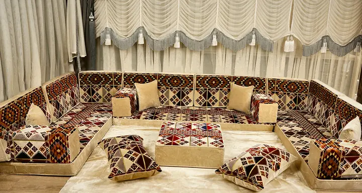 جلسة أرضية من المخمل البيج بألواح كليم بمعينات حمراء وبنية وكحلية على ثلاثة جدران مع طاولة وسط ومخدات أرضية
