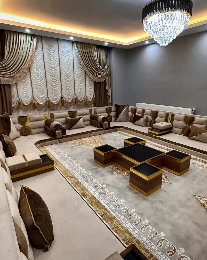 Angled view of a beige U-shaped majlis with brown armrest pads and pillows, a crystal chandelier and white festoon curtains