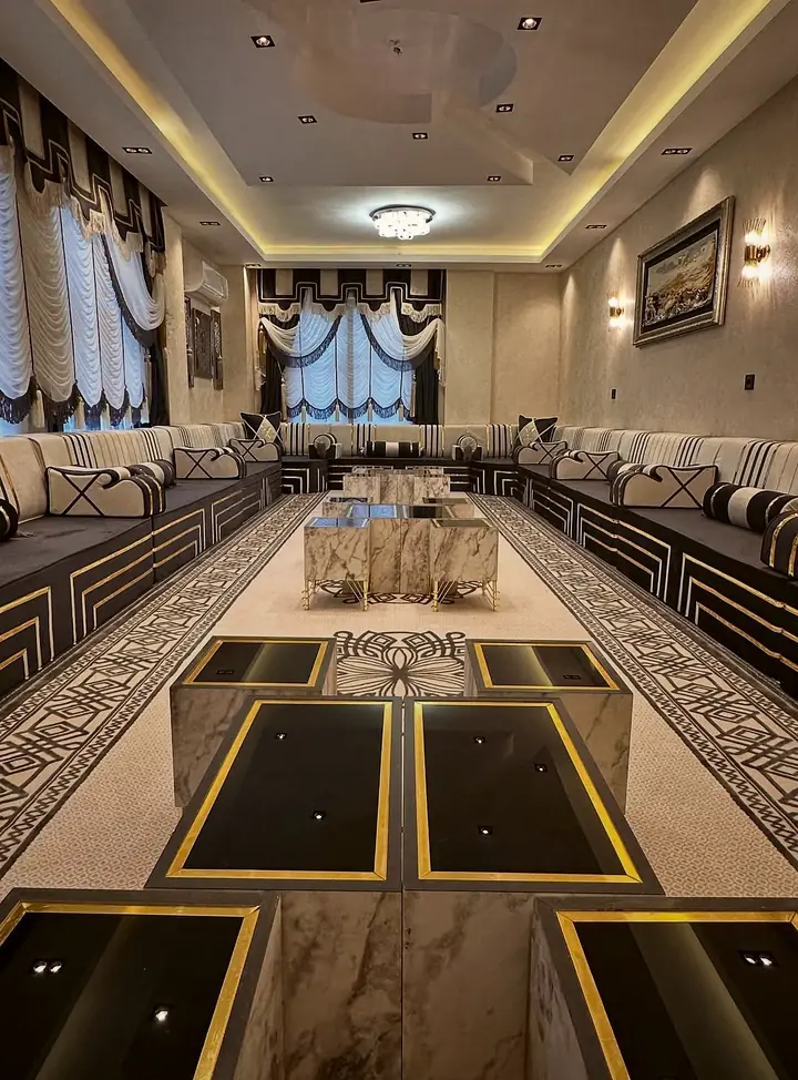 View down a hall of black and ivory majlis seating toward the window, with stepped gold lines on the bases and black-glass table tops