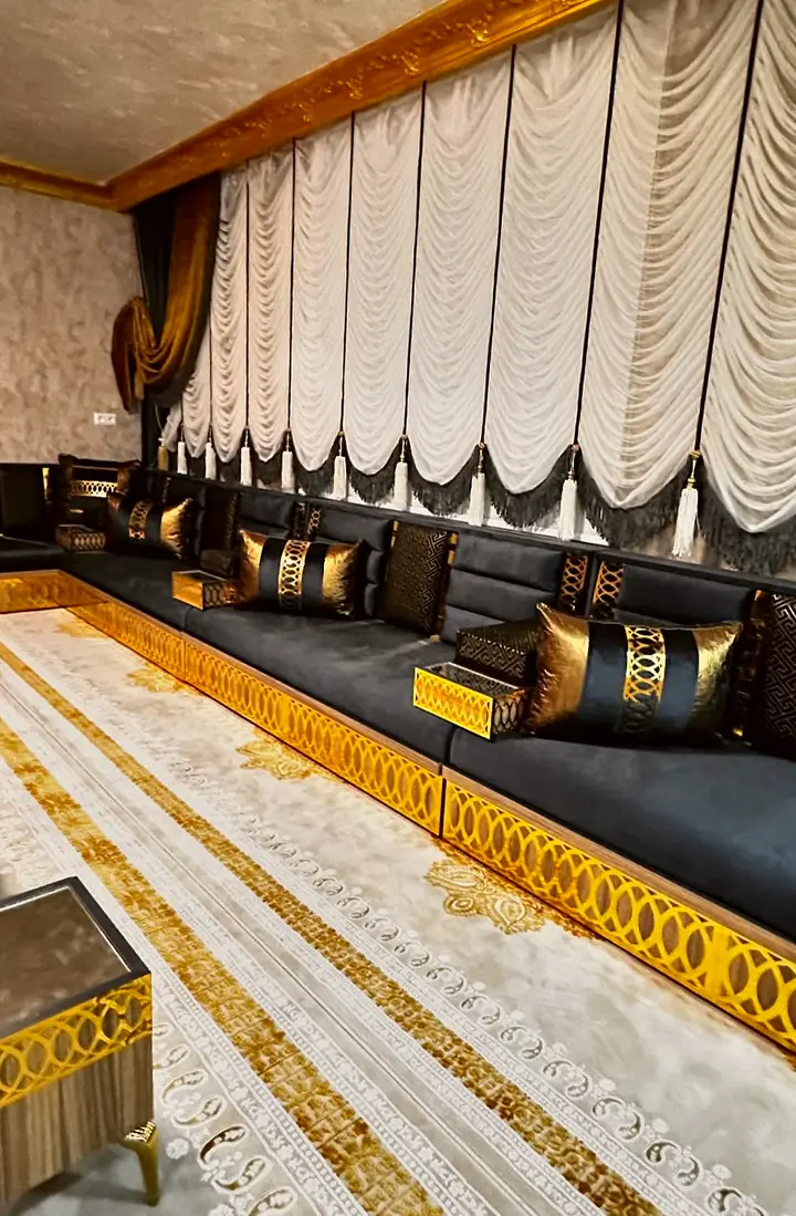 Long run of black velvet floor seating on a gold oval-lattice plinth beneath white balloon curtains, with a glass side table