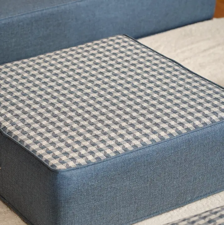 Close view of a square floor ottoman with a blue gingham top and plain blue sides, a zipper running along one side, on a white rug