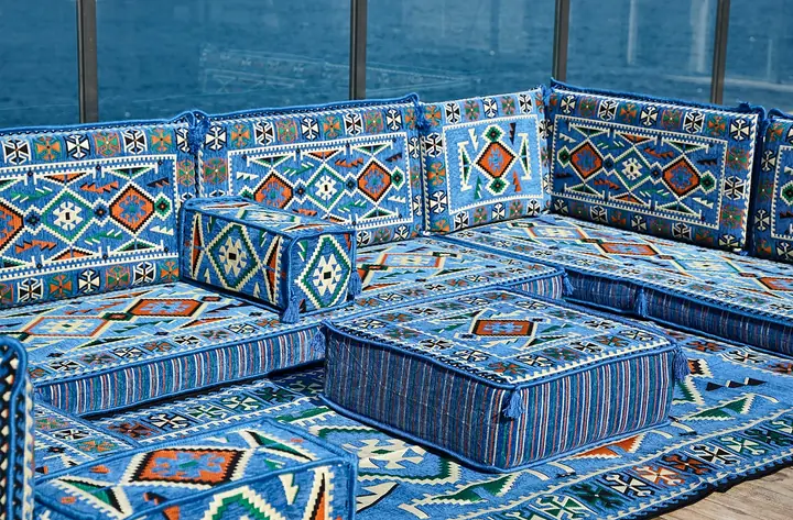 Blue kilim-pattern floor seating on three sides of a square ottoman, with striped bases, tasseled corners and a matching rug