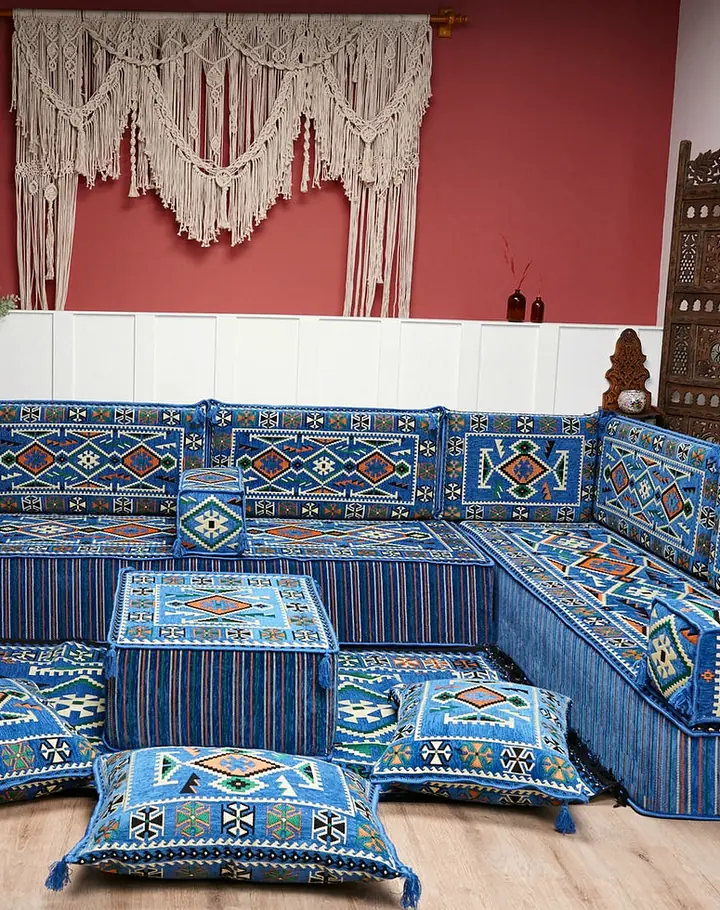 Blue kilim-pattern floor seating on three sides of a red-walled room, with an ottoman, floor pillows and carved wooden screens