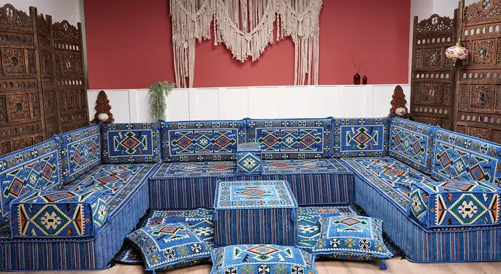 Blue kilim-pattern floor seating on three sides of a red-walled room, with an ottoman, floor pillows and carved wooden screens