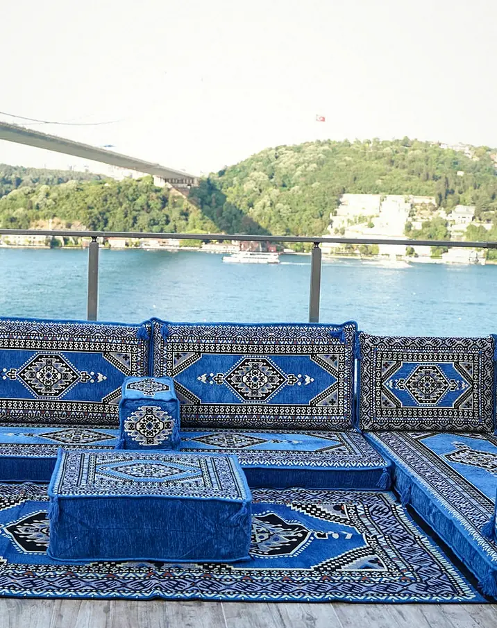 Blue medallion-pattern U-shaped floor sofa with a matching ottoman and rug on a waterside terrace, a suspension bridge behind
