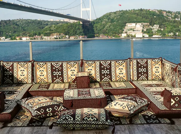 Brown and cream kilim floor sofa arranged in a U around an ottoman and floor pillows on a terrace below a suspension bridge