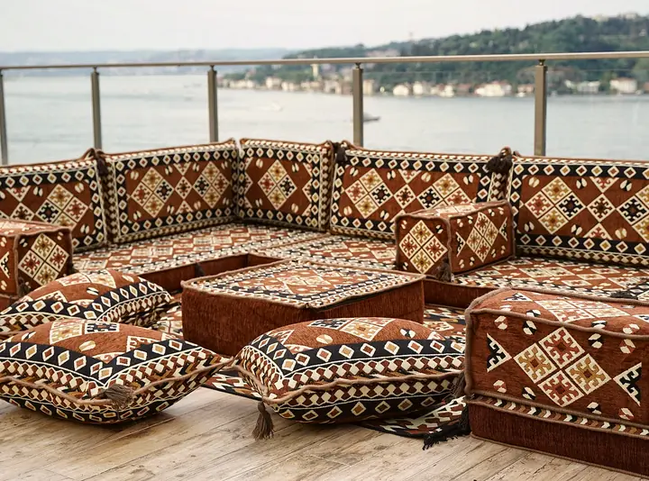 Brown U-shaped floor sofa set with cream and black diamond kilim patterns, a square ottoman and tasseled floor pillows on a wooden terrace