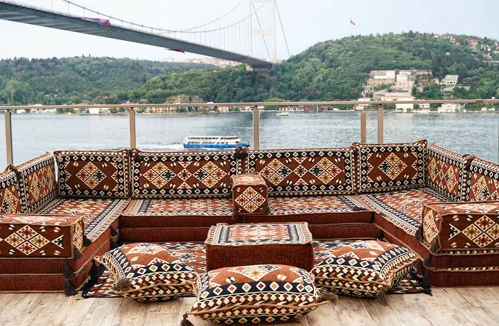 Brown and cream diamond-pattern U-shaped floor sofa set with a square ottoman and three floor pillows on a terrace above a waterway