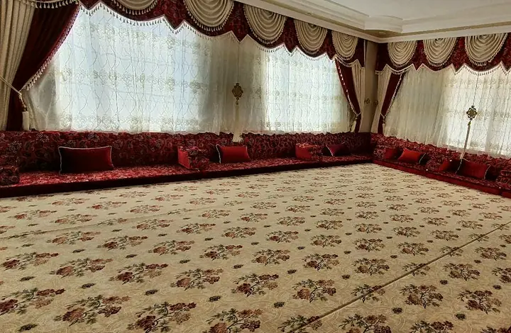 Burgundy damask floor majlis wrapped around a bright room, with red velvet pillows and a cream carpet patterned in rose sprays