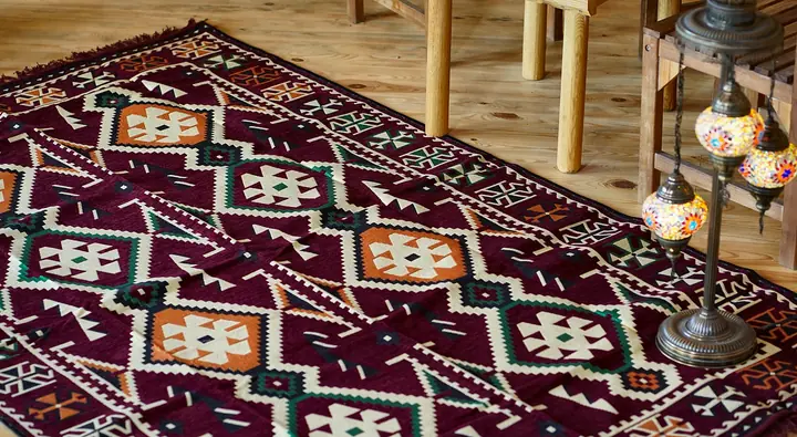 Burgundy flat-woven kilim rug with cream and orange geometric medallions on a pine floor, beside the turned legs of a wooden table