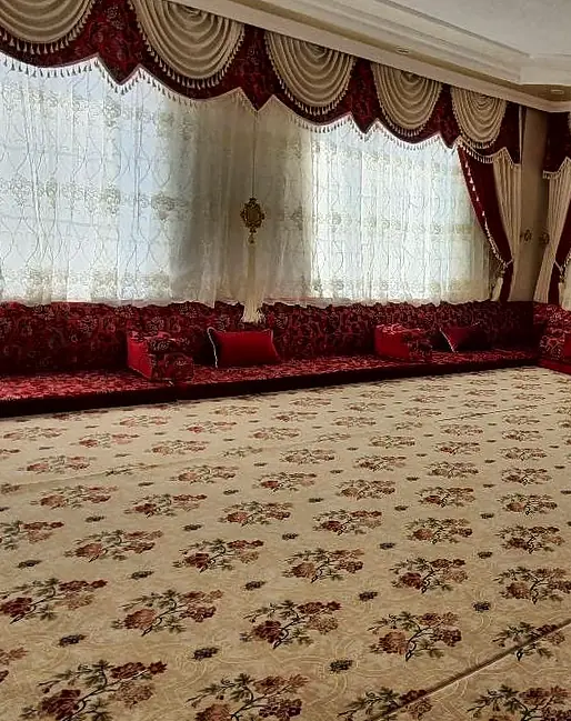 Large room with burgundy floral majlis seating along the walls, a cream floral carpet and swag valances over embroidered sheers