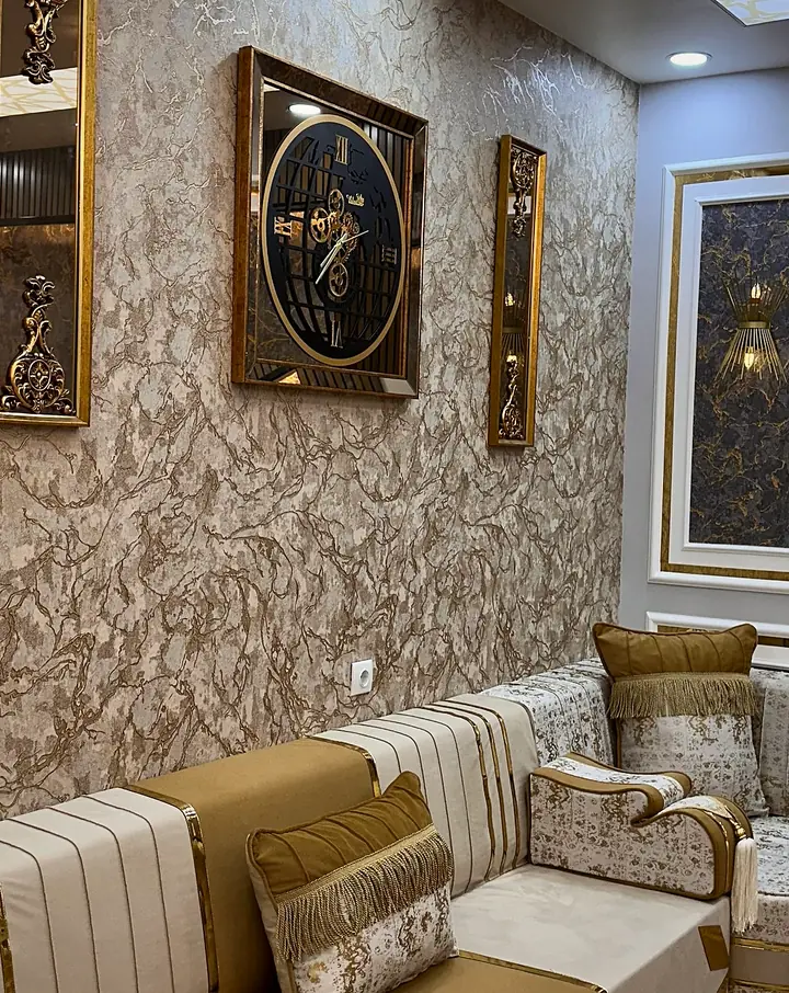Corner of a caramel and cream majlis with gold-banded cream backrests and a fringed pillow, below a wall clock and mirrored frames