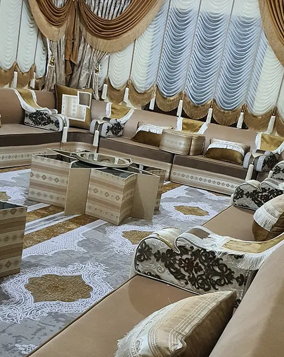 Champagne and brown majlis along the walls of a long room with gold swag curtains, a star-shaped coffee table and a gray medallion rug