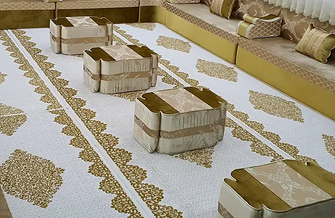Champagne and gold jacquard majlis along three walls, with four cushioned cube ottomans on a white and gold rug and tied-back sheer curtains