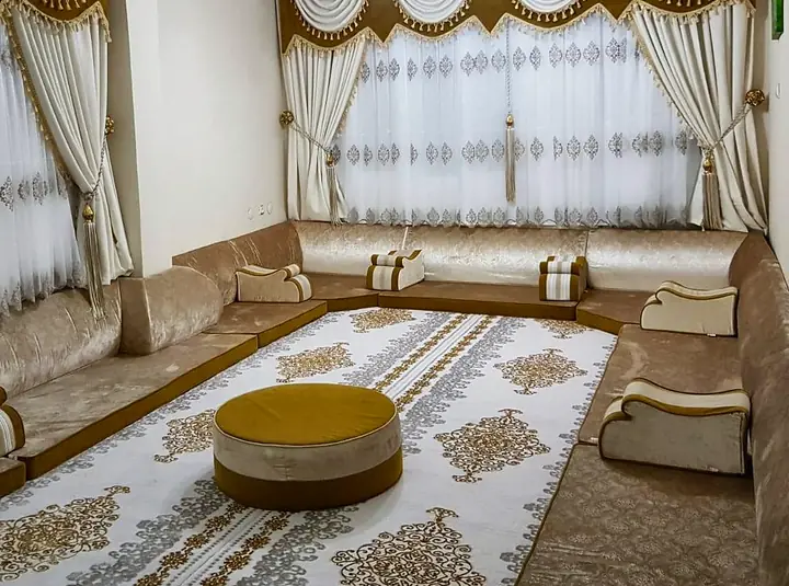 Champagne velvet floor majlis lining three walls under draped gold valances, with a mustard round pouf on a cream and gold rug