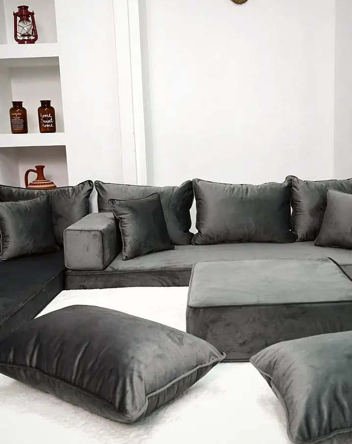 Charcoal gray velvet L-shaped floor sofa with a square ottoman and three floor pillows on a white rug in a bright room with niche shelves