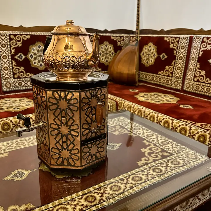 Copper samovar on a glass-topped table in front of a red and cream carved wooden bench, with a long-necked lute leaning in the corner
