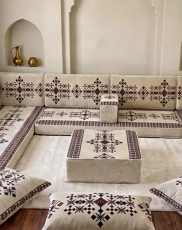 Cream velvet U-shaped floor majlis with burgundy cross-stitch motifs, a square ottoman and floor pillows below brass vessels in niches