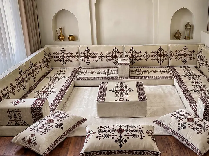 Cream velvet U-shaped floor majlis with burgundy cross-stitch motifs, a square ottoman and floor pillows below brass vessels in niches