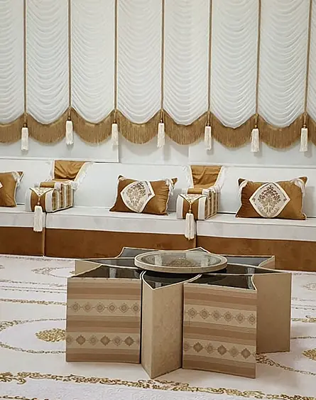 Cream majlis on caramel velvet bases along three walls with star-shaped coffee tables, a gold-patterned cream rug and white swag blinds