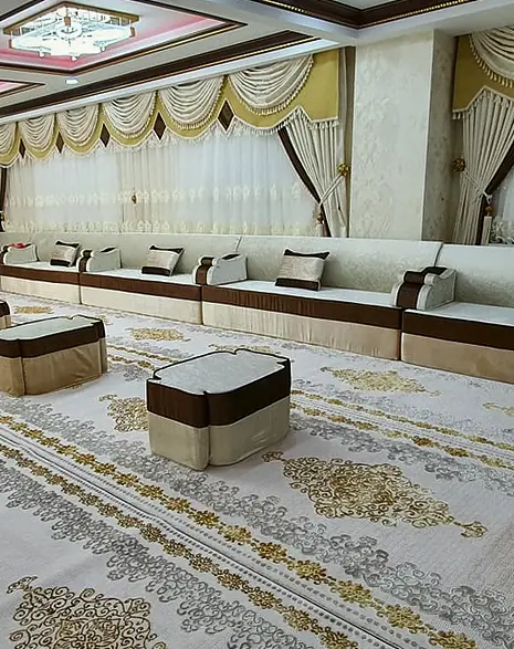 Cream and chocolate majlis along the walls of a large hall, with banded poufs on a gold and cream carpet and layered window curtains