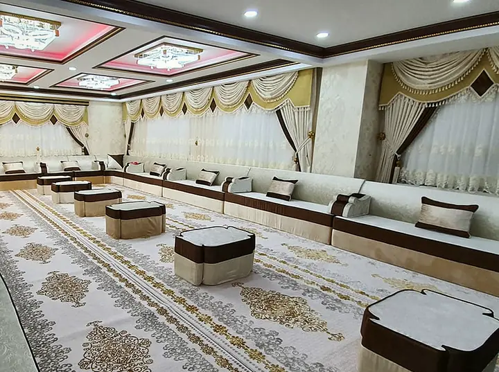 Long reception hall lined on every wall with cream majlis seating trimmed in chocolate brown, low poufs set down a gold-patterned carpet