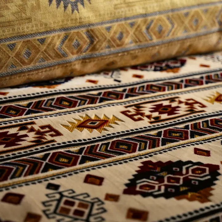 Close view of cream kilim fabric with maroon, black and gold geometric bands running under a gold patterned cushion edge
