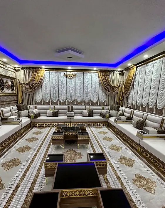 Cream majlis around a room lit by a blue ceiling cove, with gold-trimmed curtains and walnut tables topped in black glass