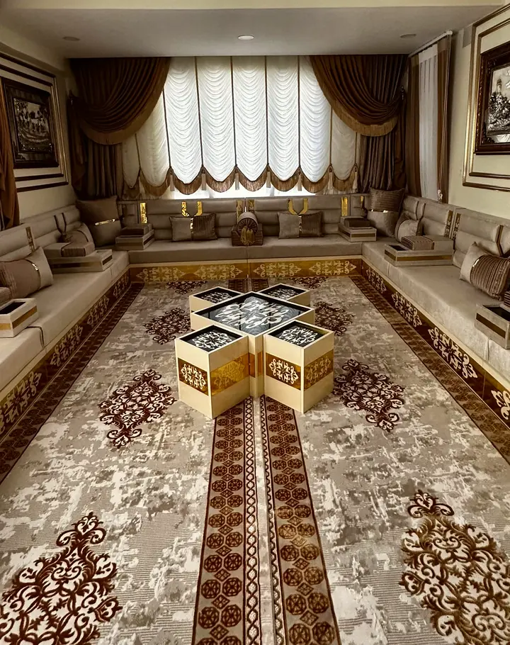 Cream majlis seating around a long room with gold box tables grouped on a brown medallion carpet below gathered ivory blinds