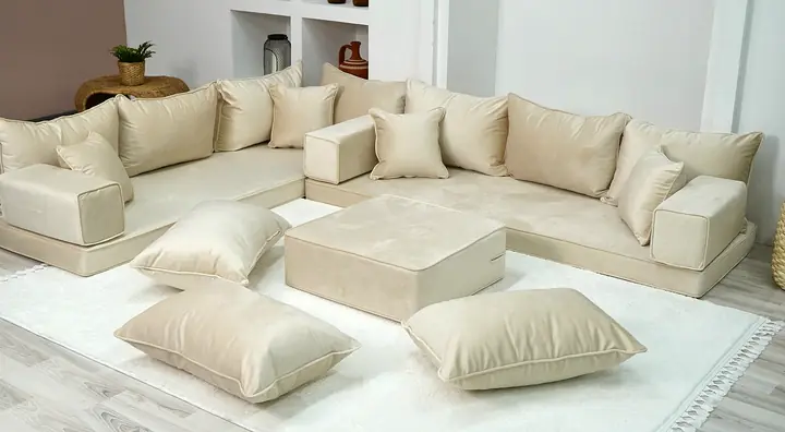 Cream floor sofa arranged in an L with matching backrests, a square ottoman and three floor pillows on a pale rug