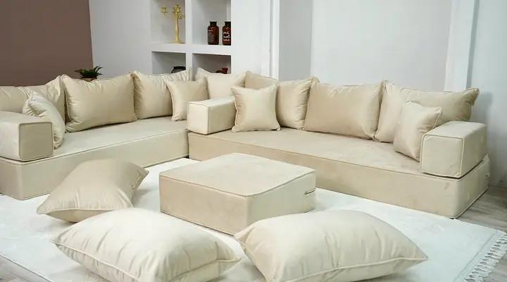 Cream velvet L-shaped floor sofa with a matching ottoman and floor pillows arranged on a white rug in a light room