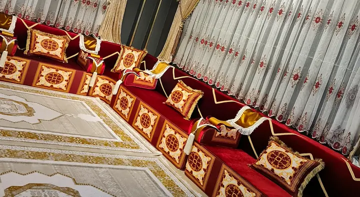 Crimson velvet majlis with ivory and gold medallion panels along two walls, a cream rug and white embroidered curtains with red valances