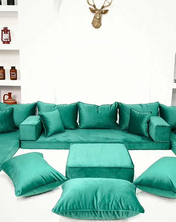 Emerald green velvet U-shaped floor sofa with a square ottoman and three floor pillows on a white rug in a white room with shelves