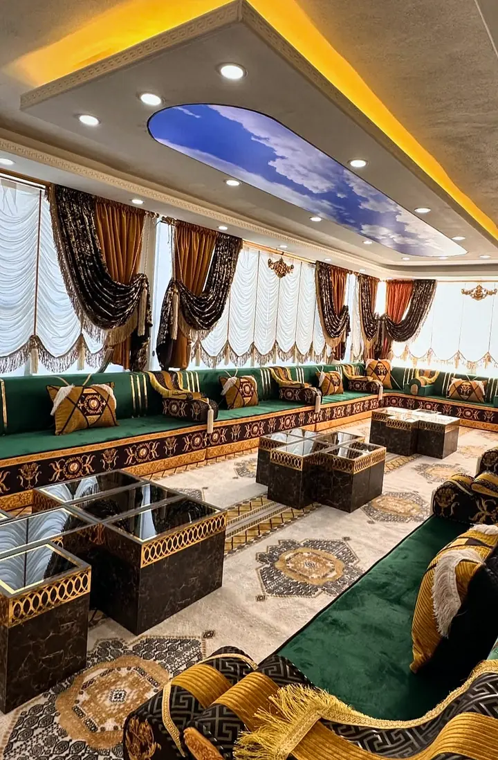 Fringed black-and-gold armrest bolster in front of a U of green seating around glass-topped tables under a sky-painted ceiling