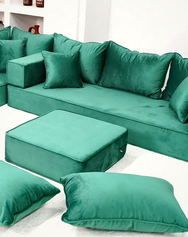 Emerald velvet L-shaped floor sofa with thick seats, bolster armrests and back pillows, an ottoman and three floor pillows on a white rug