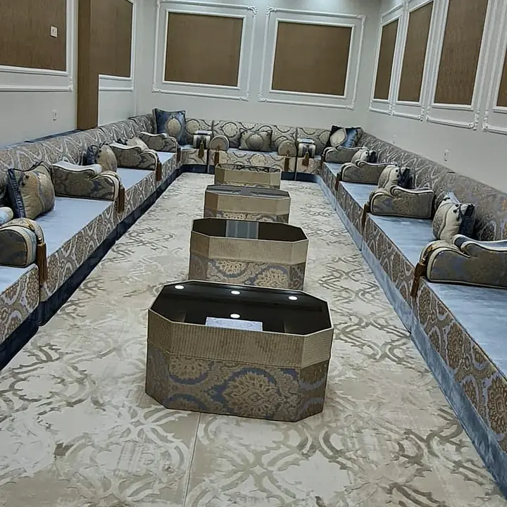 Long hall with blue-grey velvet majlis seating along both walls, gold jacquard armrests and octagonal tables down the middle