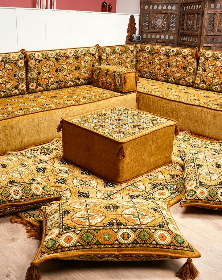 Gold Afghan-pattern L-shaped floor sofa with a square ottoman and four tasseled floor pillows beside a carved wooden screen
