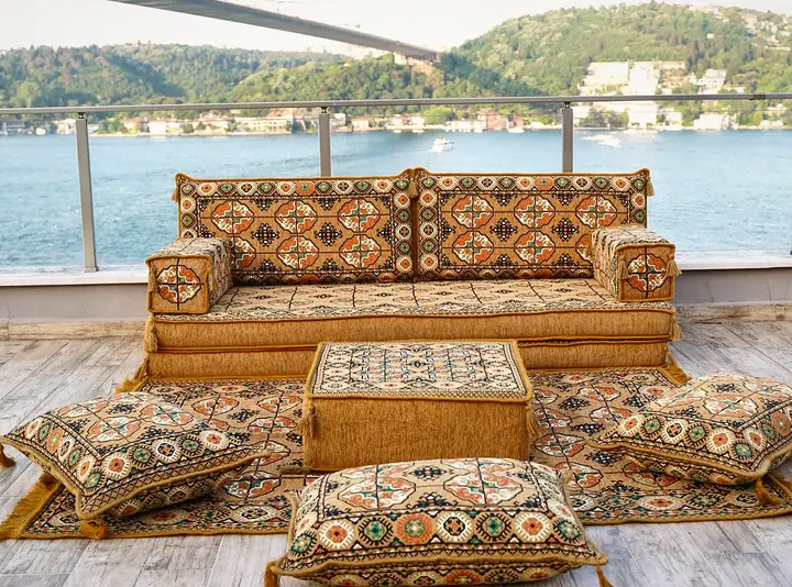 Gold single floor sofa set in a medallion-pattern weave with an ottoman, floor pillows and a fringed rug on a terrace above the water