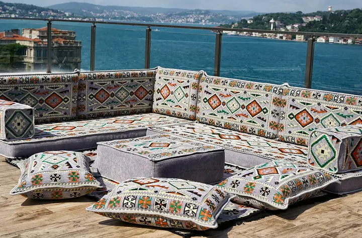Gray kilim L-shaped floor sofa set with a square ottoman, bolster armrests and three patterned floor pillows on a sunny terrace