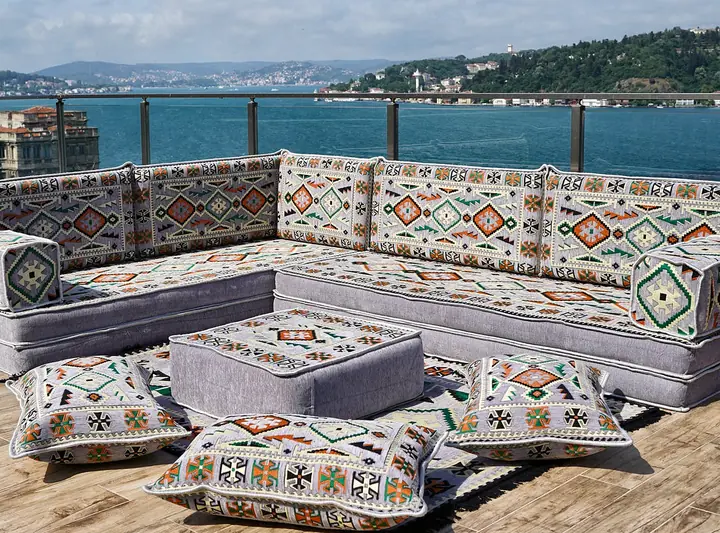 Gray L-shaped floor sofa with kilim-pattern backrests, bolster armrests, a square ottoman and three floor pillows on a waterside terrace
