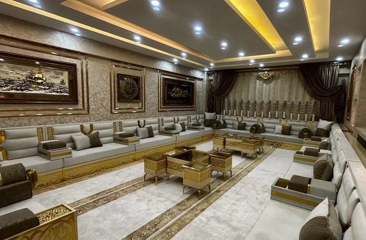 Gray and white majlis on three walls with gold cross straps, gold armrest panels and a cluster of gold-framed low tables