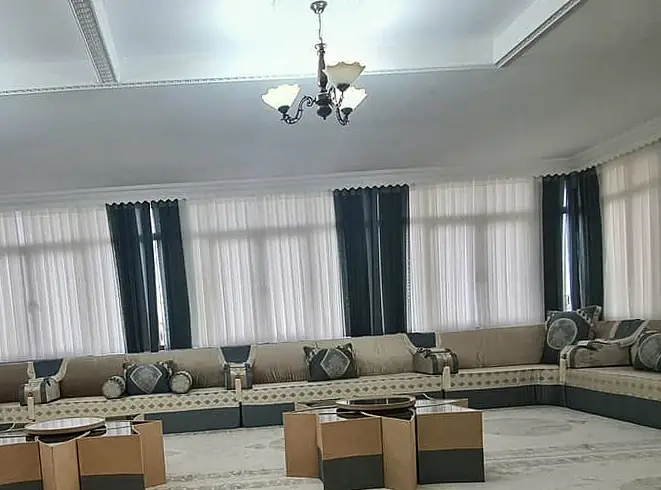 Very large hall ringed by beige majlis on dark green bases under a single chandelier, with sheer curtains and nesting cube tables