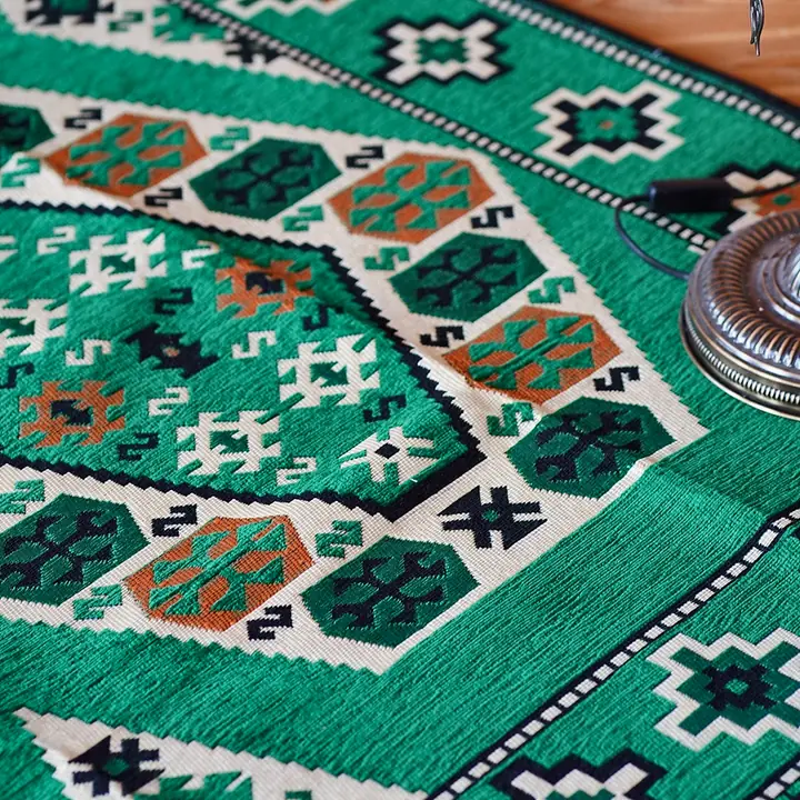 Close view of a green rug with cream, orange and black motifs on the floor, the brass base of a floor lamp at its edge