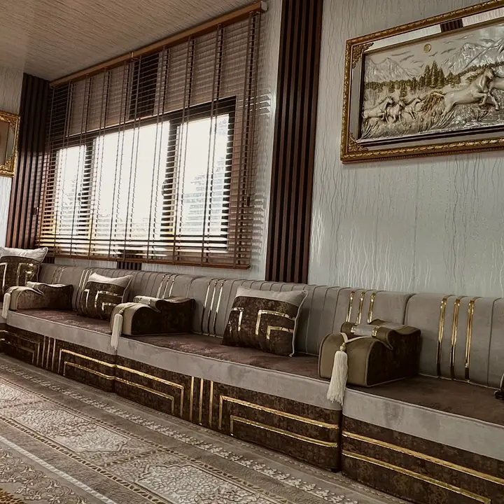 Long greige and brown seating run beneath a wide window and framed relief art, with gold Greek-key pillows along it